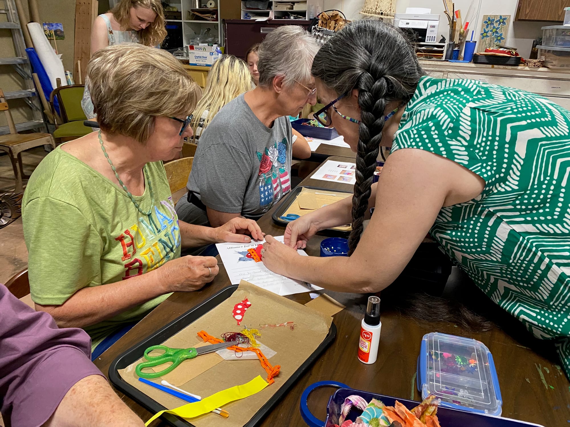 Woman in braids reaches towards another woman's artwork to assist with her collage project.